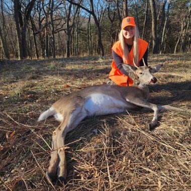 A young woman poses with a spike buck harvested while deer hunting.