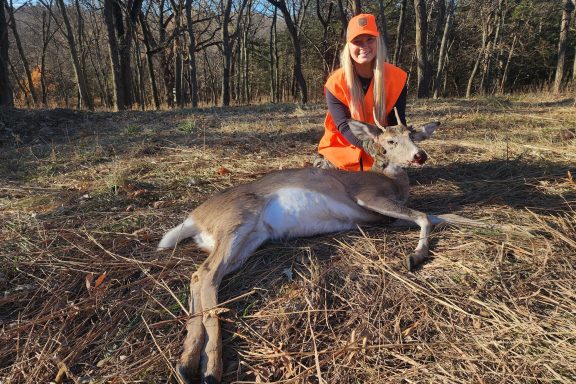 A young woman poses with a spike buck harvested while deer hunting.