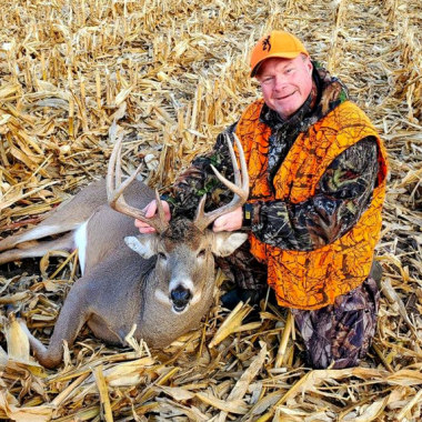 A man in hunter orange poses alongside a harvested buck in a stubble corn field.