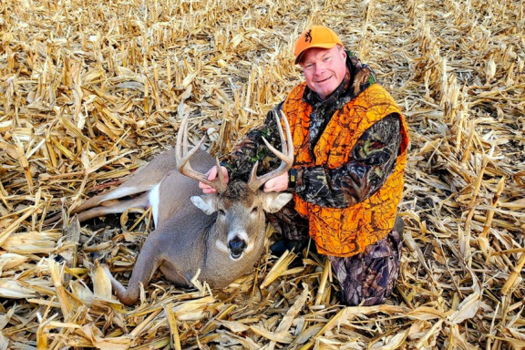 A man in hunter orange poses alongside a harvested buck in a stubble corn field.