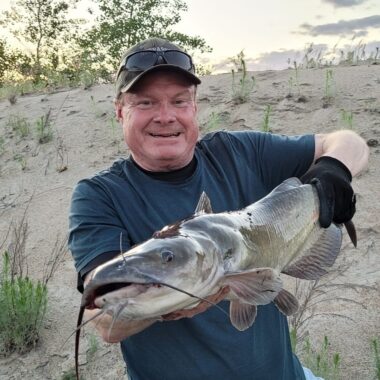 Greg Wagner holds a large channel catfish on a sandy shoreline.