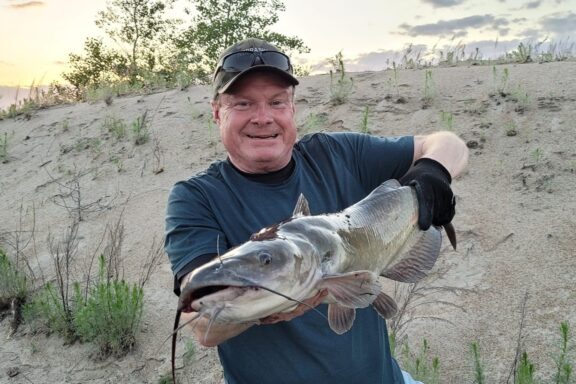 Greg Wagner holds a large channel catfish on a sandy shoreline.
