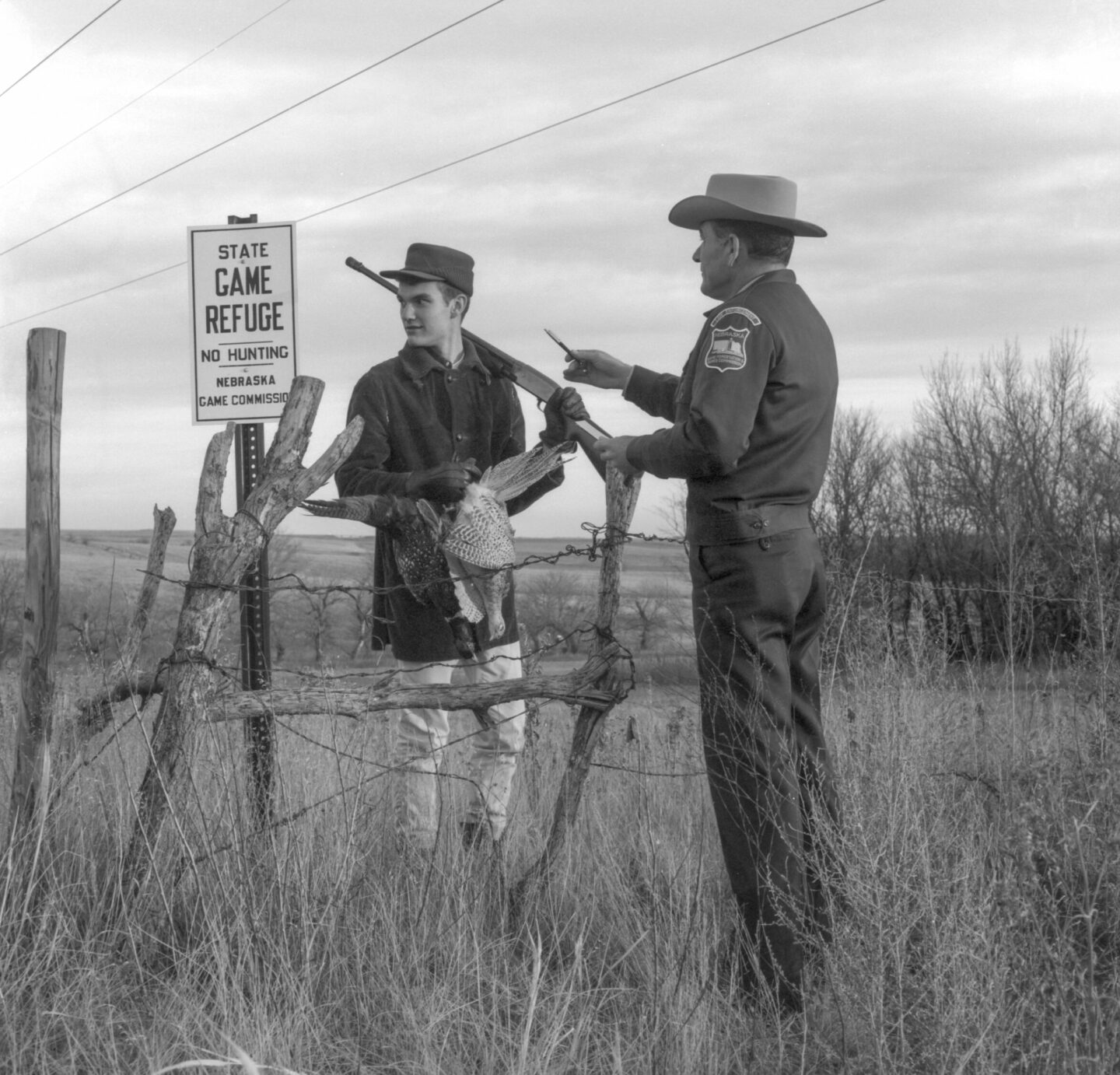 Law Enforcement Infraction Shots. "Youthful Violator." Conservation officer Carl Gettmann points out "no hunting" sign on game refuge to teenage boy carrying firearm and bagged pheasants. [Black and White Negative 2306.] Photographer unattributed, undated. [Before Feb. 1967.] Copyright Nebraskaland Magazine/Nebraska Game and Parks Commission.
