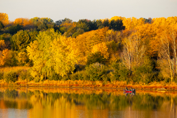 Anglers fish from a boat during fall at a tree-lined lake.